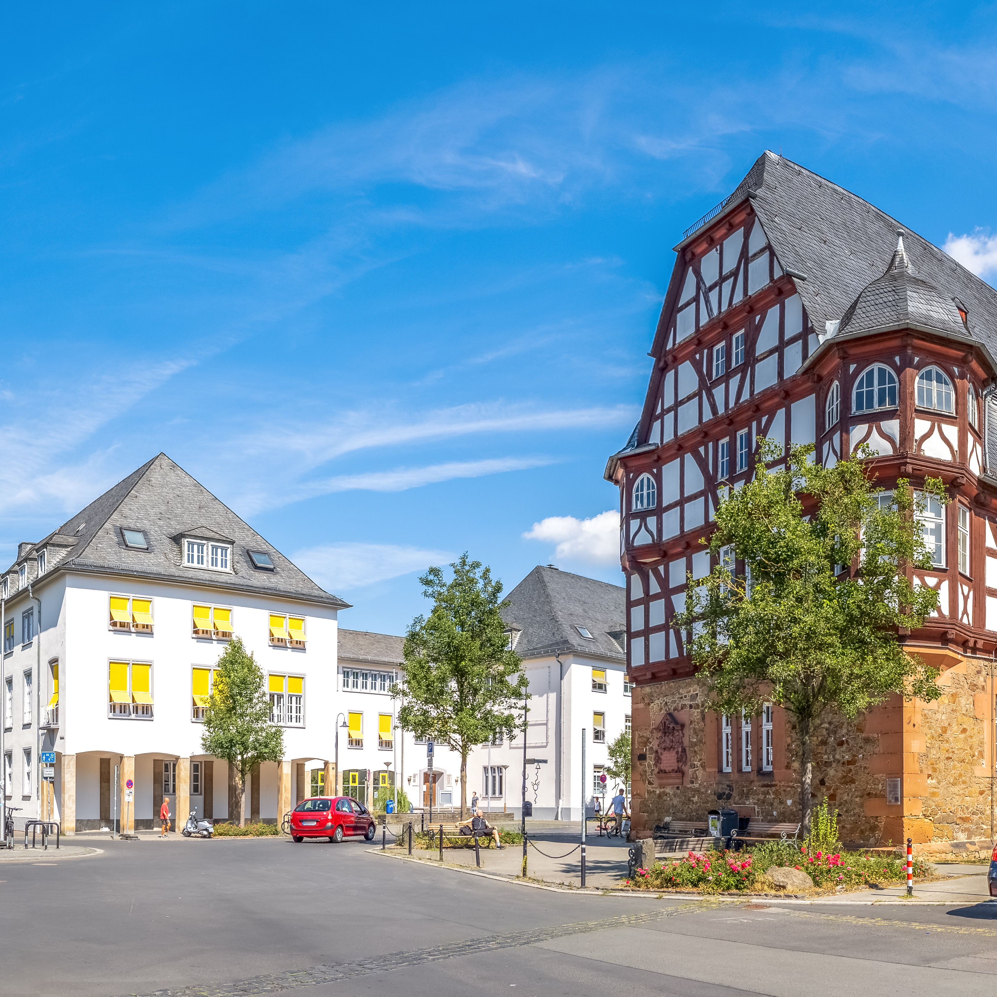 Großes Fachwerkhaus und andere Gebäude, umgeben von Bäumen und parkenden Autos bei Sonnenschein.