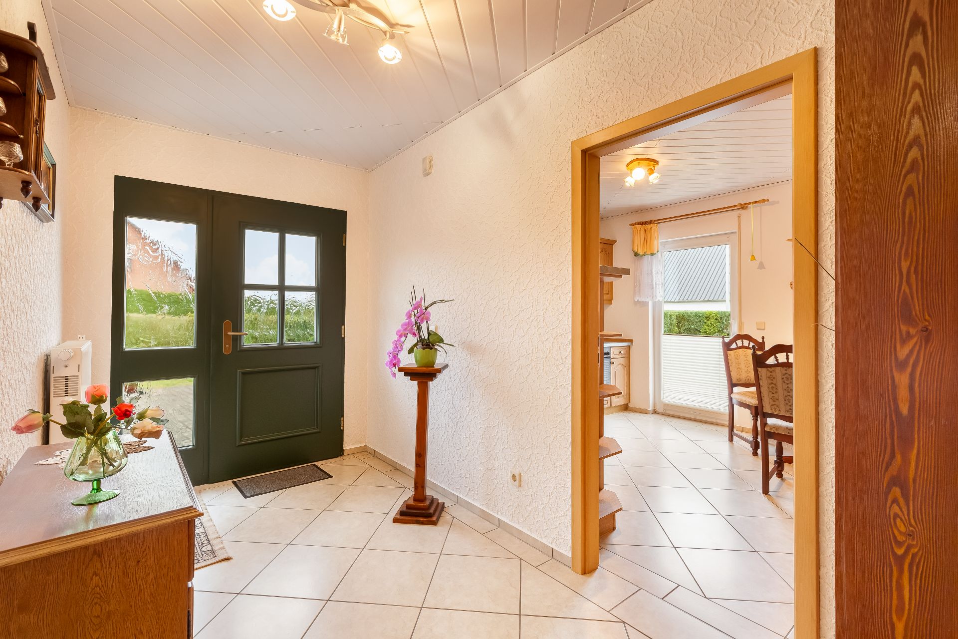 Heller Flur mit dunkler Haustür, Kommode, Blumen und Blick in ein Esszimmer mit Stühlen.