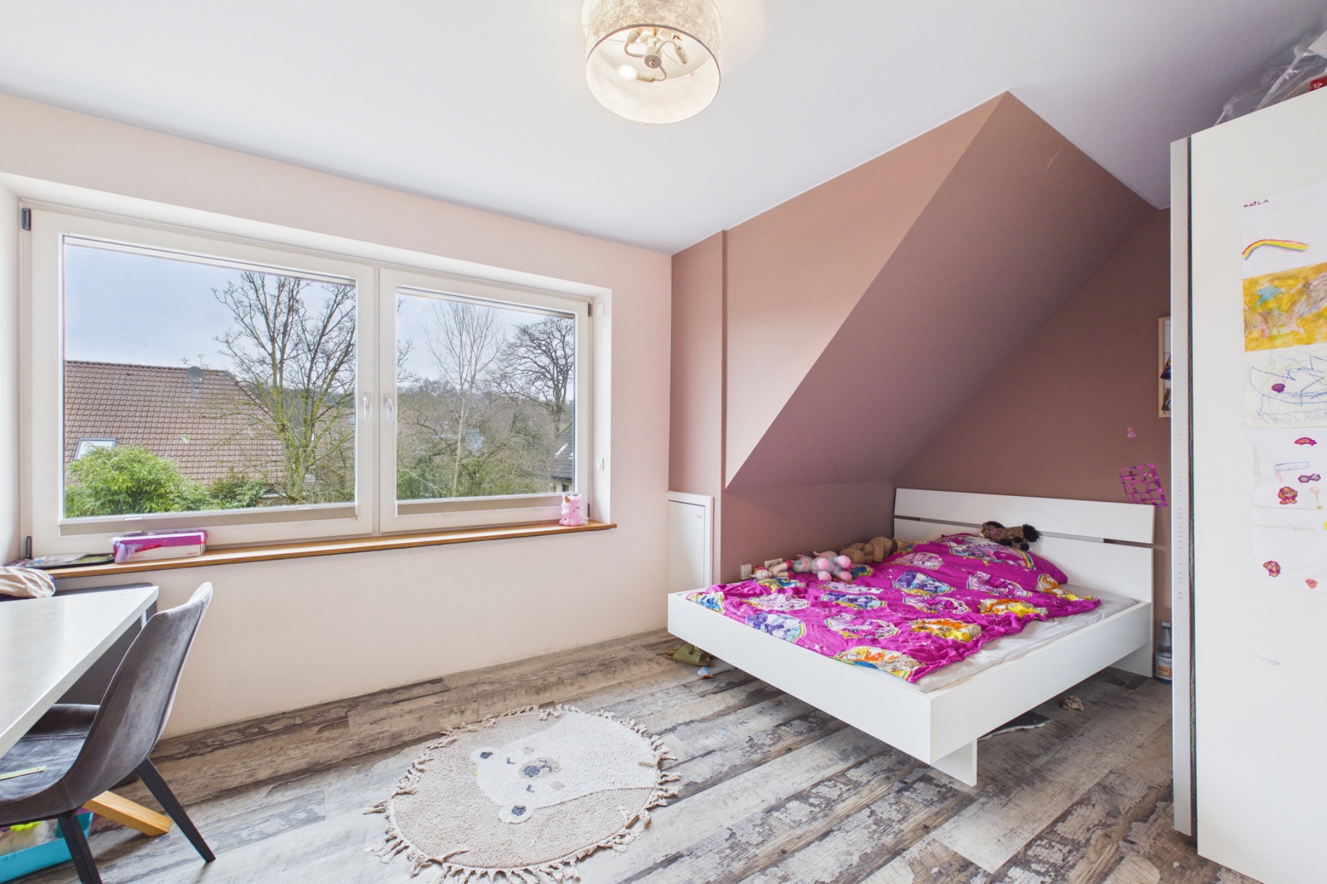 Helles Kinderzimmer mit Einzelbett, buntem Bettzeug, Schreibtisch und großem Fenster.