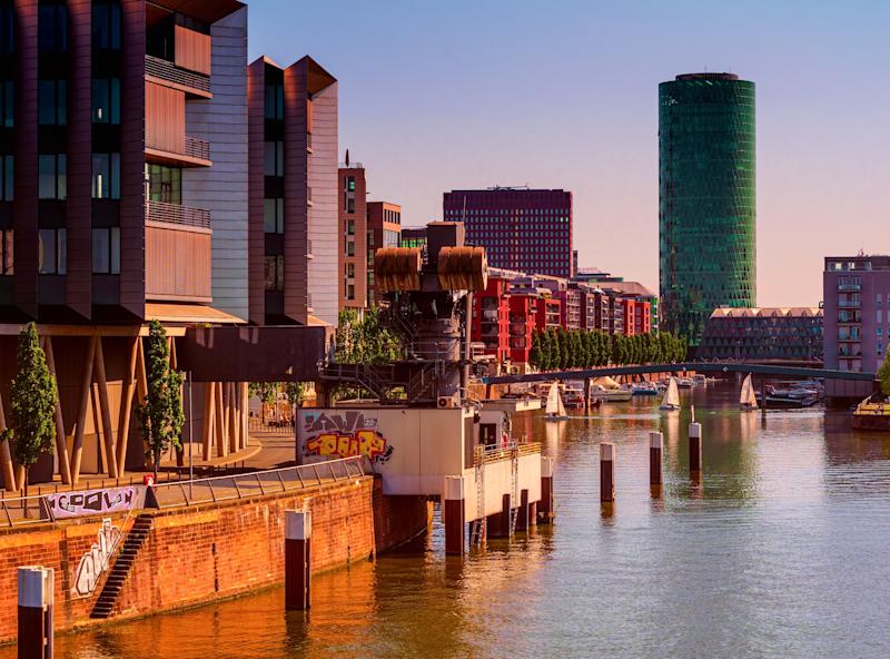 Moderne Wohngebäude am Flussufer mit Booten und Hochhäusern im Hintergrund.