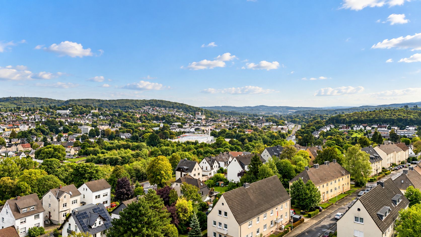 Blick auf eine Wohnsiedlung mit Häusern, Bäumen und hügeliger Landschaft im Hintergrund.