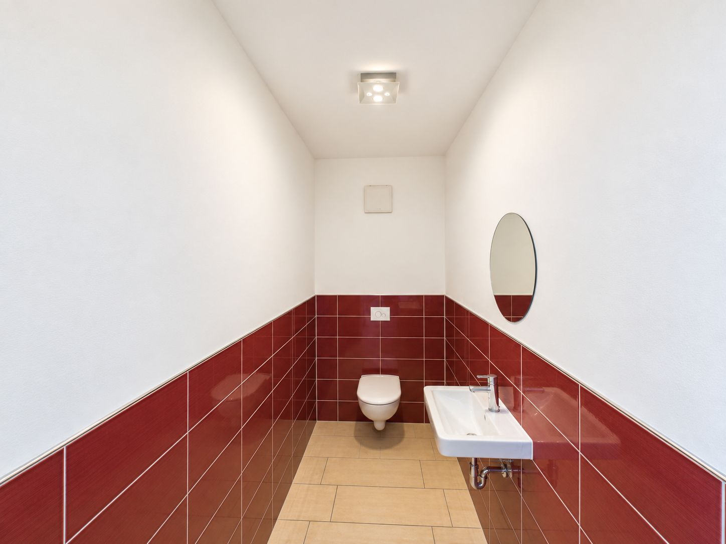 Schmales Bad mit Wandtoilette, kleinem Waschbecken und rotem Fliesenspiegel.