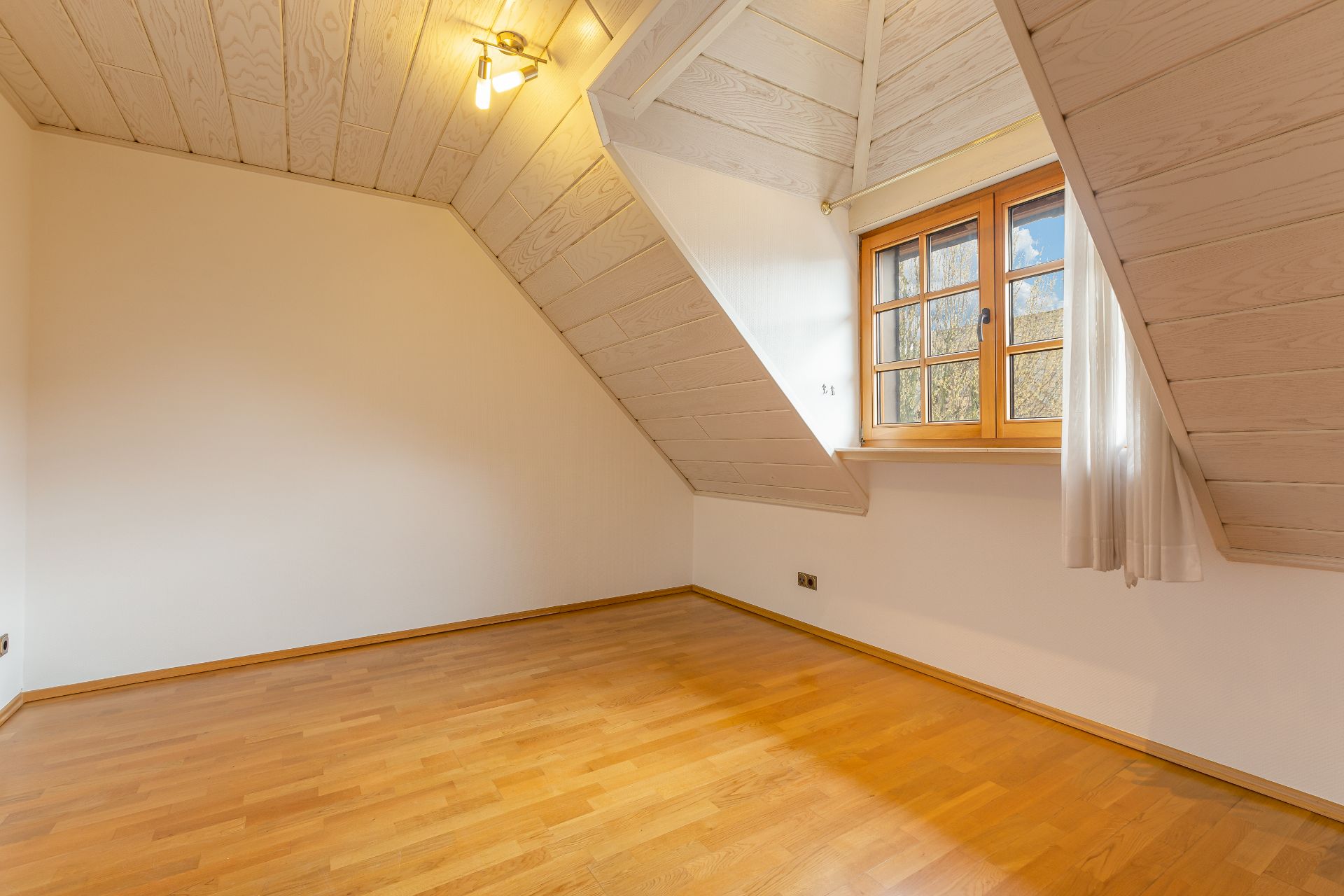Leeres Zimmer mit Holzfußboden, Dachschräge und Fenster mit Gardine.