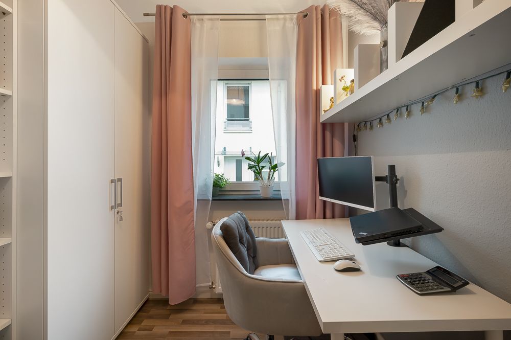 Kleines Arbeitszimmer mit Schreibtisch, Monitor, Tastatur, grauem Stuhl und Fenster mit rosa Vorhängen.