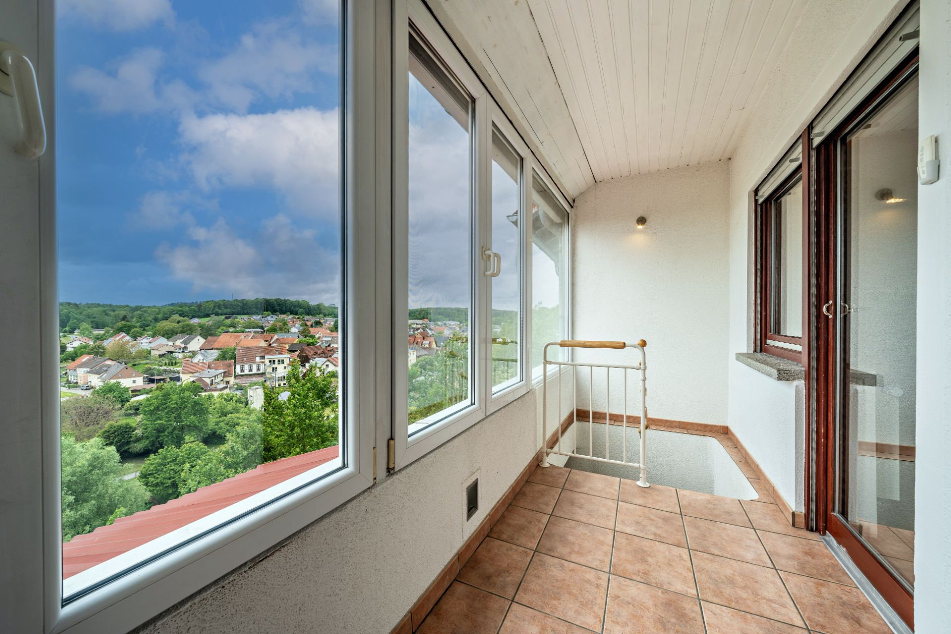 Veranda mit großen Fenstern, Fliesenboden und Blick auf grüne Landschaft und Häuser.