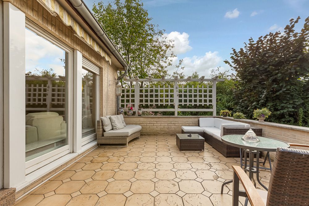 Sonnige Terrasse mit Sitzmöbeln, Tisch und Blick auf Garten, angrenzend an Wohnhaus mit großen Fenstern.