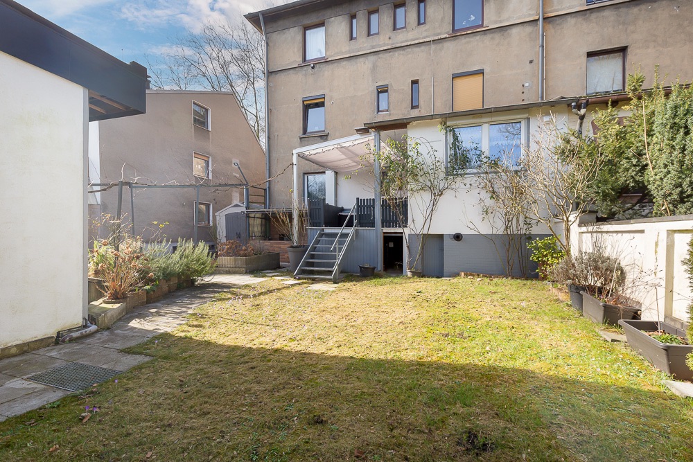 Garten mit Rasenfläche, Terrasse und Treppe hinter einem mehrstöckigen Wohnhaus.