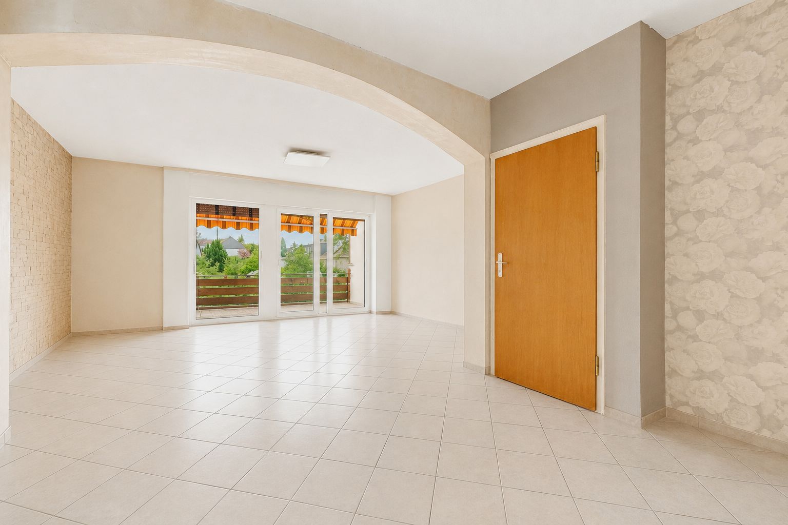 Heller, leerer Raum mit Fliesenboden, großen Fenstern und Holztür, Zugang zu Balkon mit Markise.