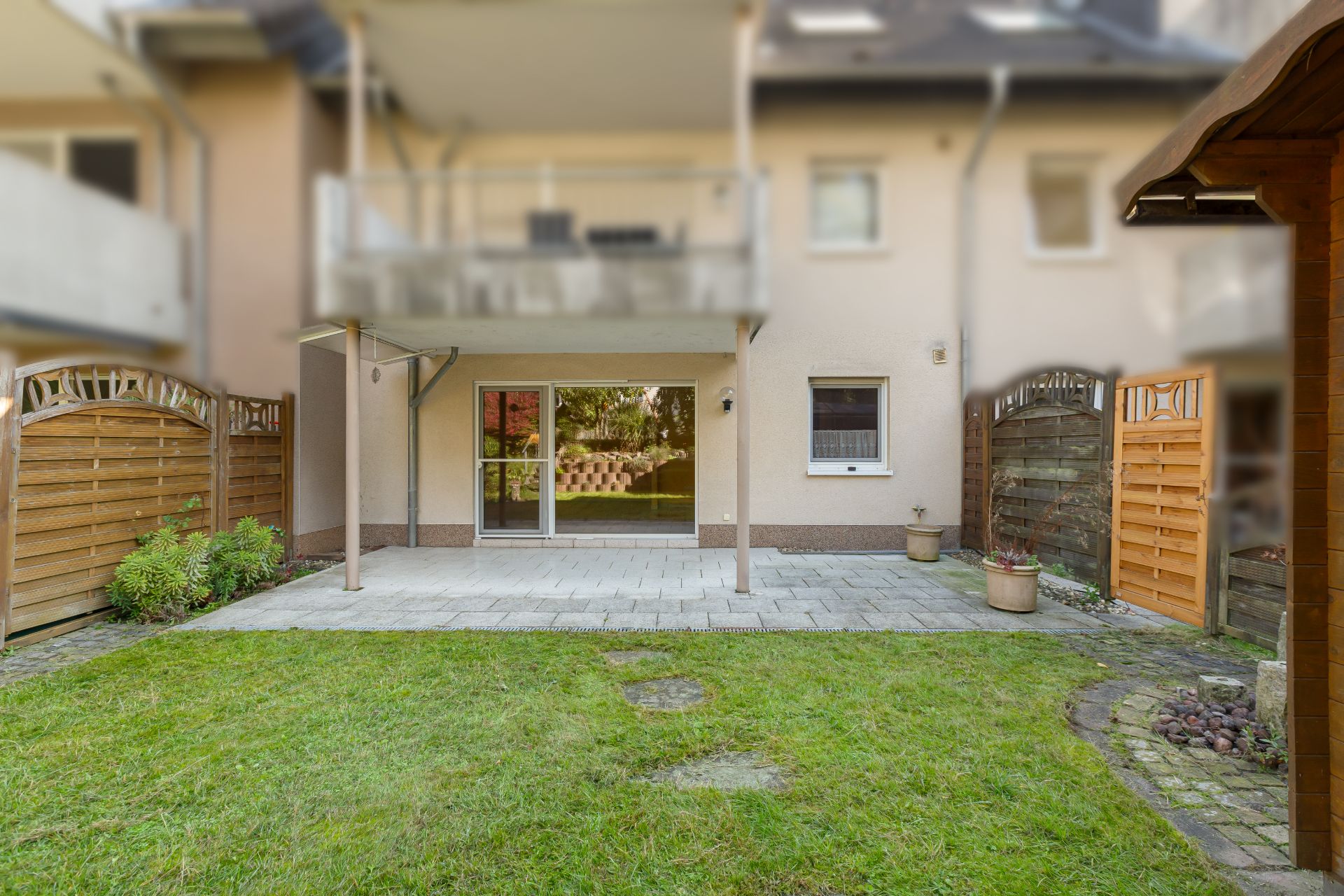 Garten mit Rasenfläche, Steinterrasse, Holzzaun und bepflanzten Blumentöpfen vor einem Wohnhaus.