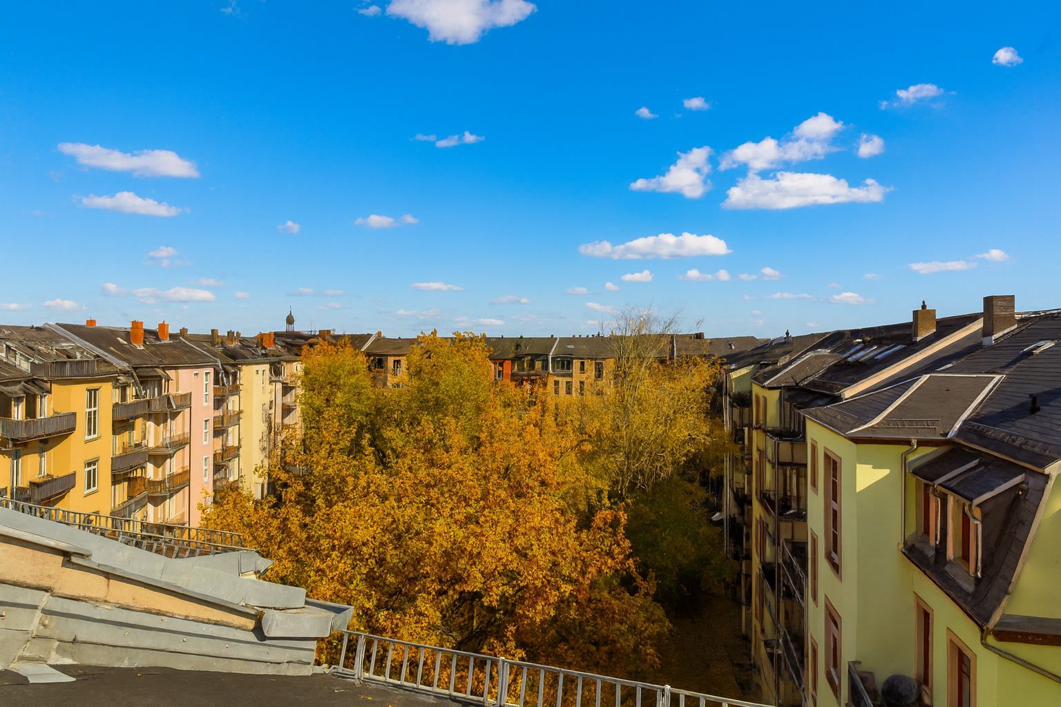 Blick von einem Balkon auf Wohnhäuser und herbstliche Bäume unter blauem Himmel.