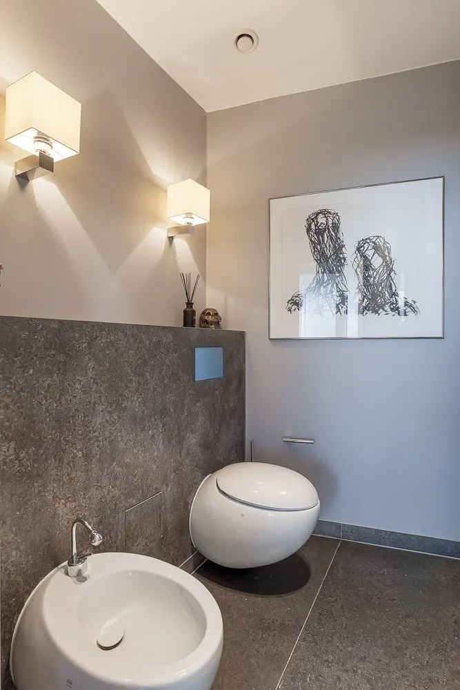 Modernes Badezimmer mit rundem WC, Bidet, Wandlampen und abstraktem Bild an der Wand.