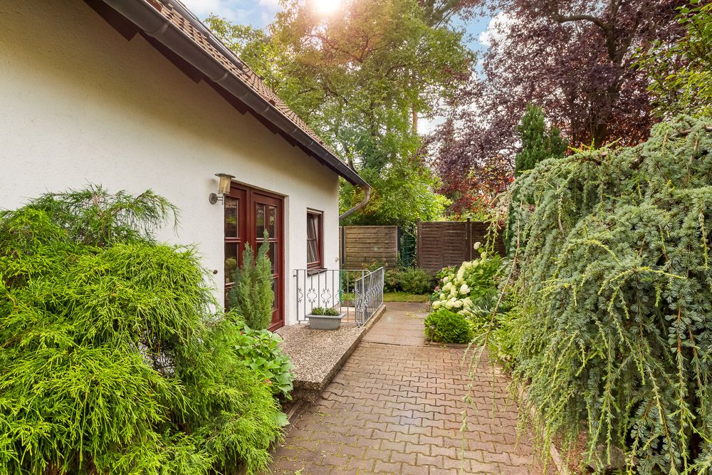 Gepflasterter Weg neben einem Haus mit Holztür, umgeben von grünen Büschen und Bäumen.