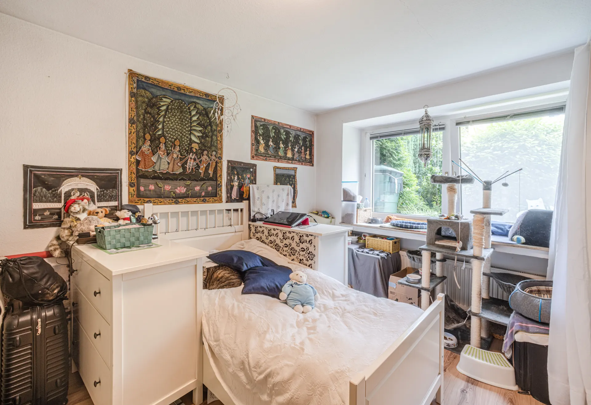 Helles Schlafzimmer mit Einzelbett, Katzenbaum, Wandteppichen und großem Fenster.