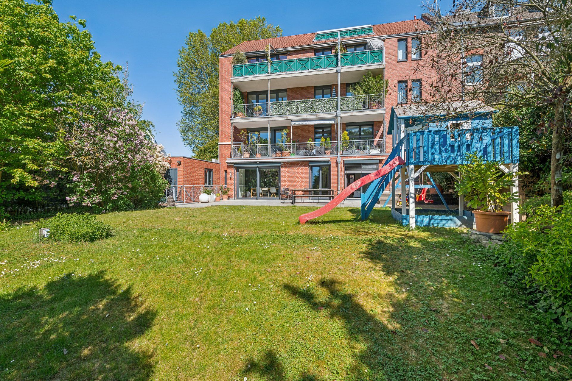 Mehrfamilienhaus mit Balkonen, großem Garten und Kinderspielplatz mit Rutsche im Vordergrund.