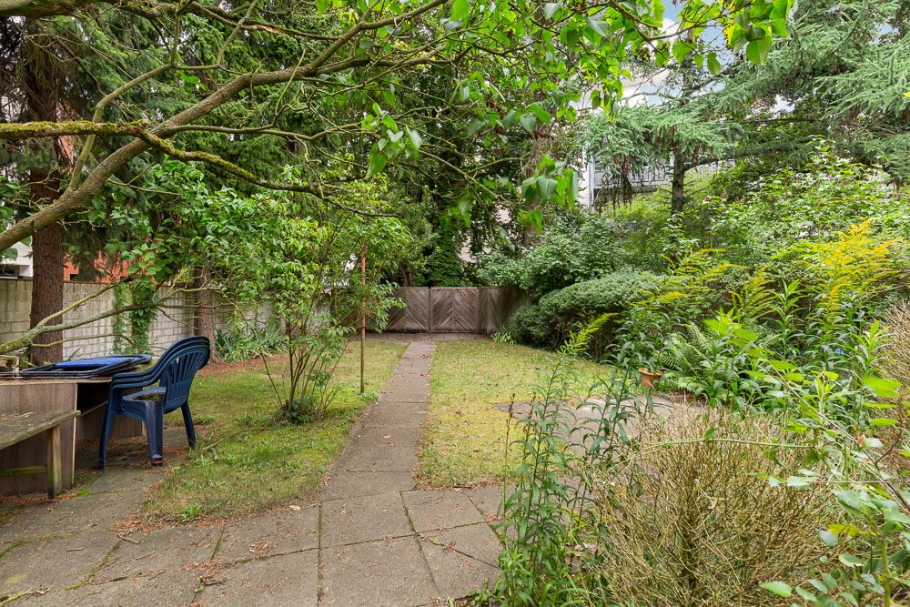 Garten mit Gehweg, Bewuchs und zwei blauen Kunststoffstühlen am Holztisch.