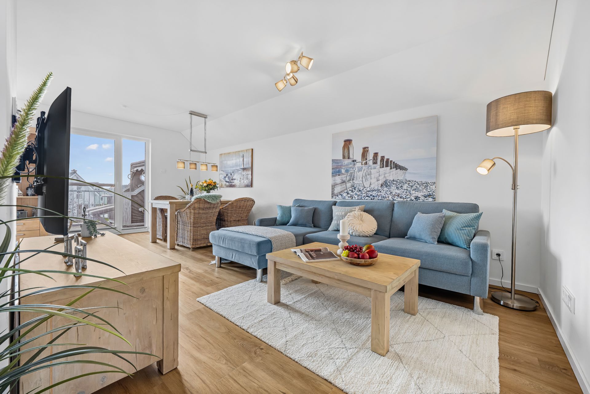Helles Wohnzimmer mit blauem Sofa, Holztisch, Fernseher und Zugang zu einem Balkon.