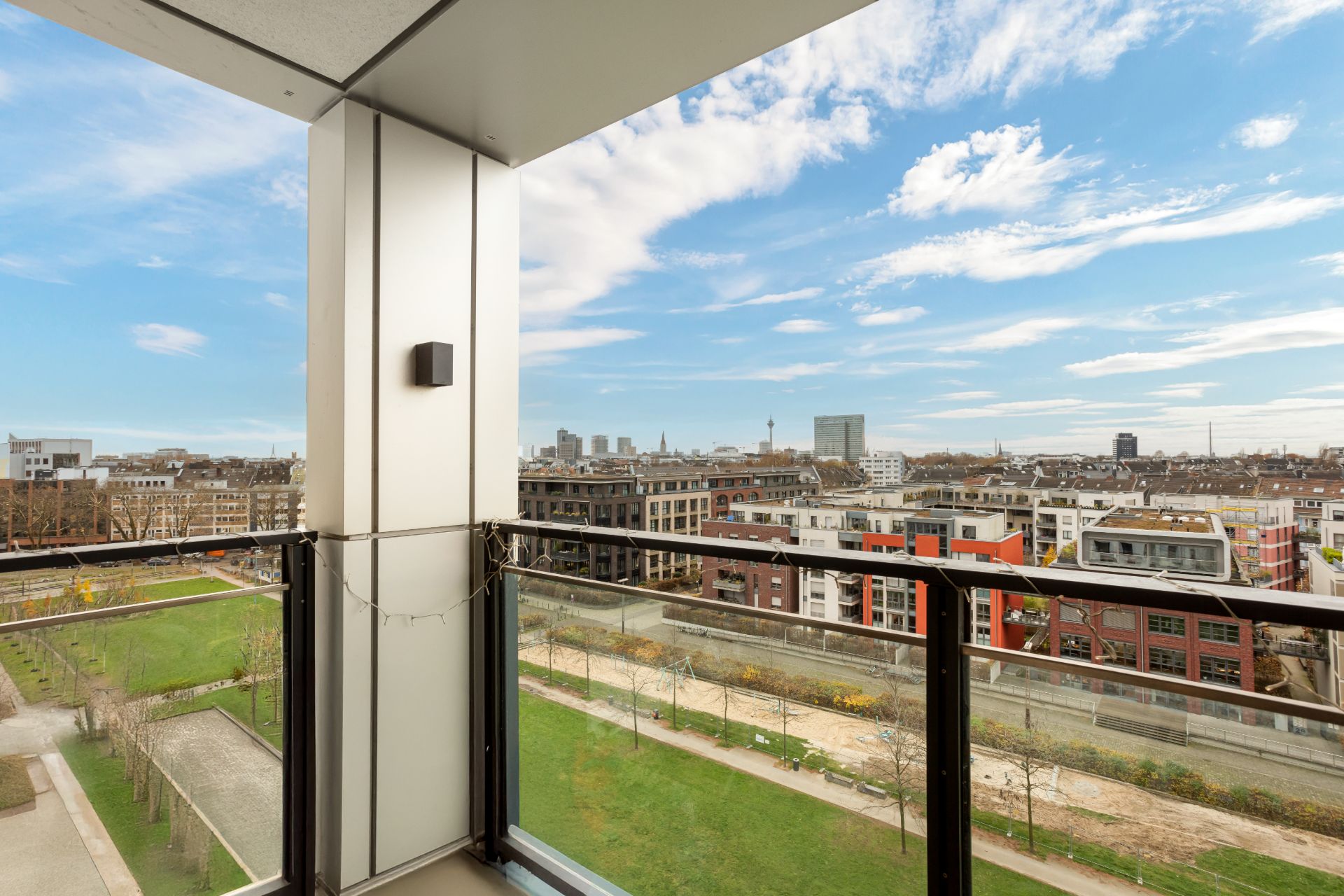 Blick von einem Balkon mit Glasgeländer auf Wohnhäuser und eine Grünfläche.