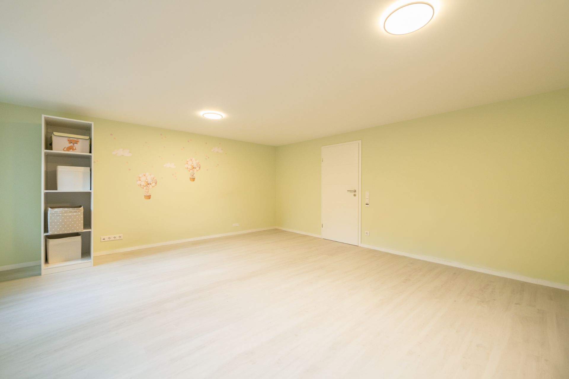Helles, leeres Zimmer mit grünlichen Wänden, weißer Tür und Regal mit Körben links.