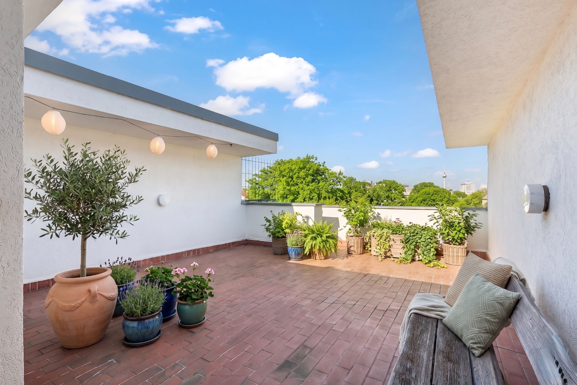Große Dachterrasse mit Pflanzen in Töpfen, Sitzbank und Lichterkette, umgeben von weißen Wänden.
