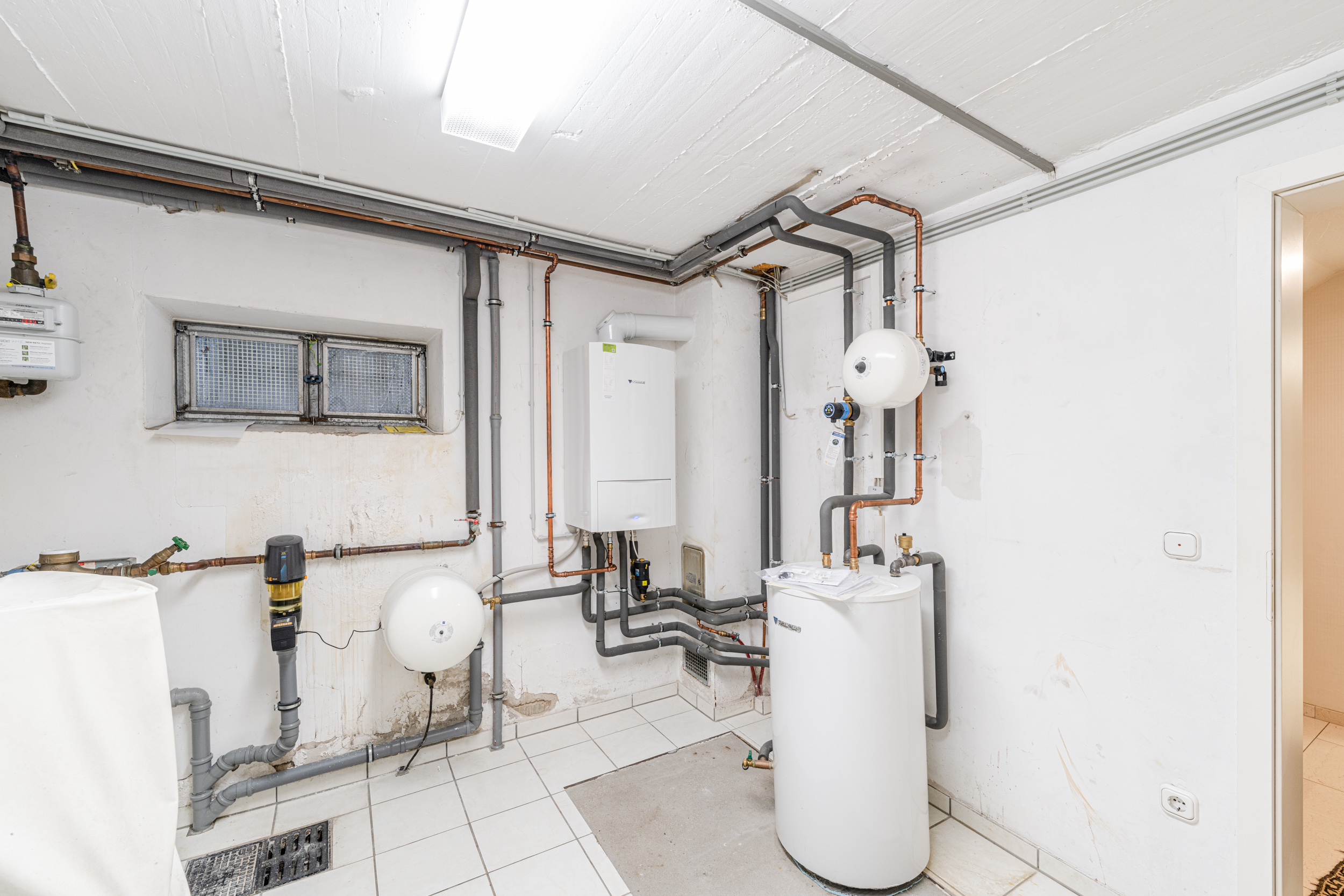 Heller Heizungsraum mit Rohren, Boiler und Wandgerät. Fenster an der Wand.