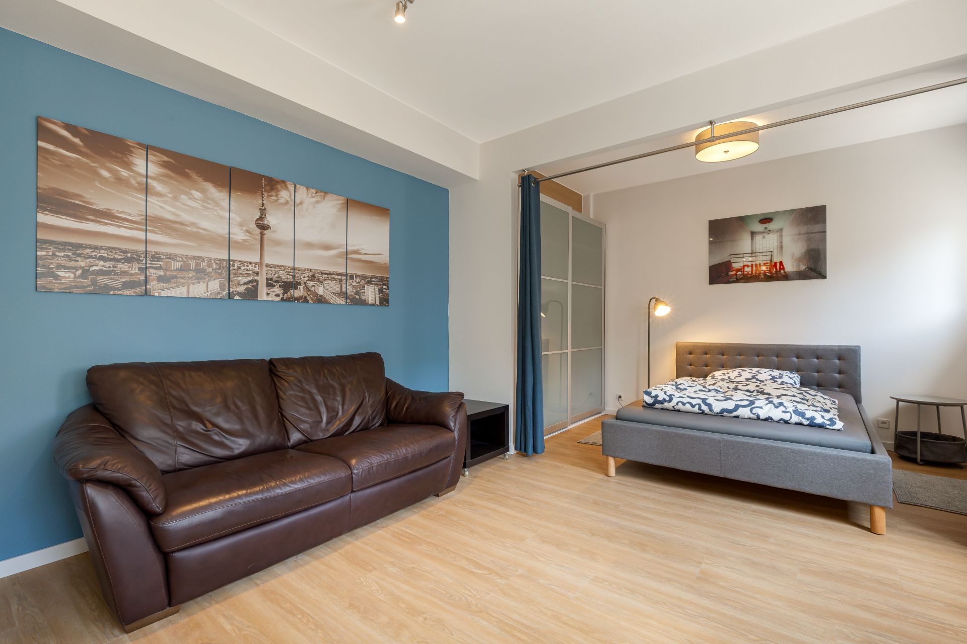 Wohnzimmer mit braunem Ledersofa, grauem Bett und blauer Wand mit Bild.