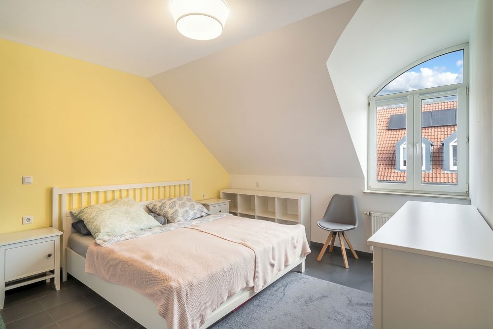 Helles Schlafzimmer mit Doppelbett, gelber Wand, Fenster, Stuhl und Kommode.