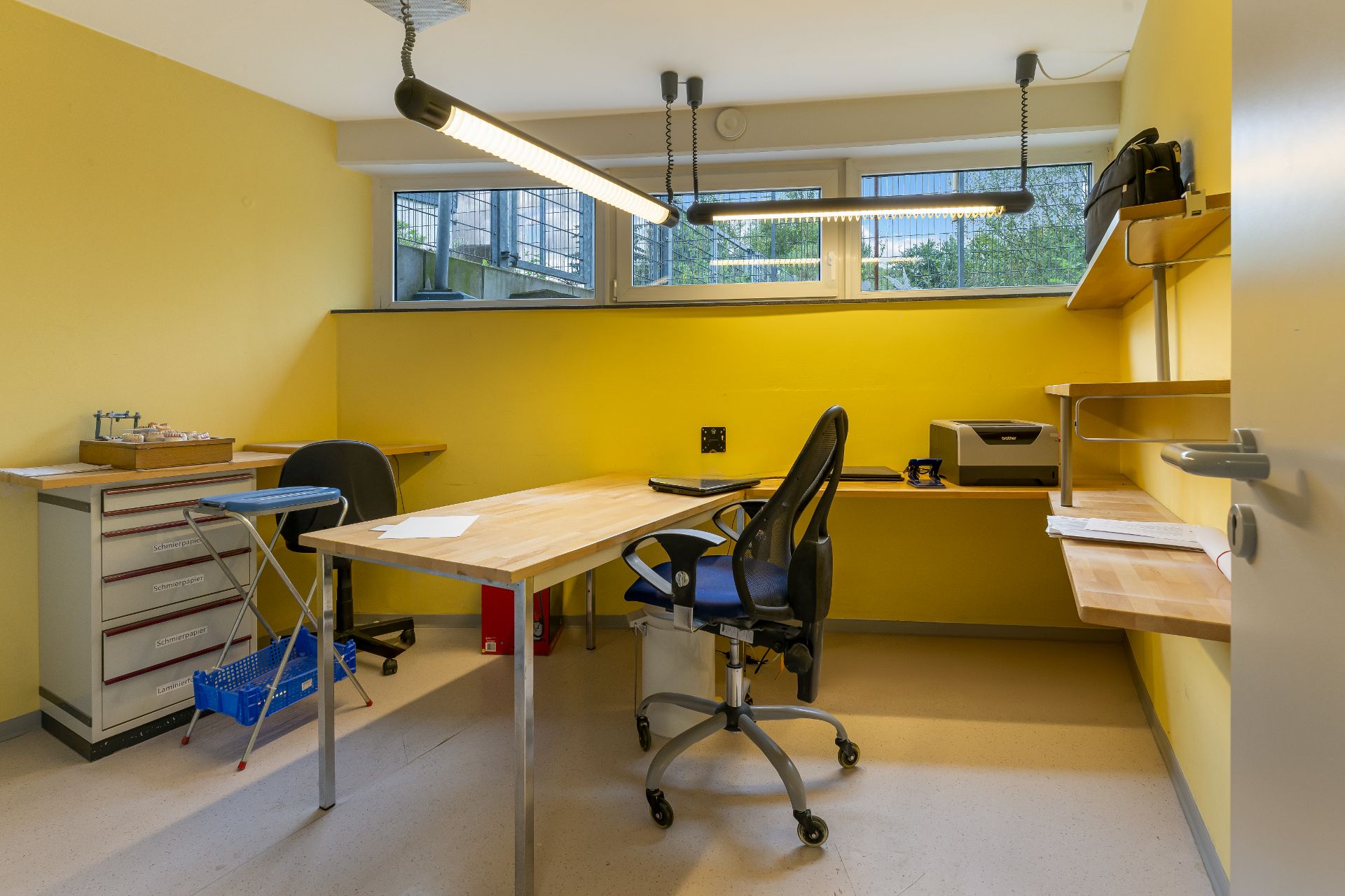 Helles Arbeitszimmer mit gelben Wänden, Schreibtisch, Stuhl, Regalen und Drucker unter Fenstern.