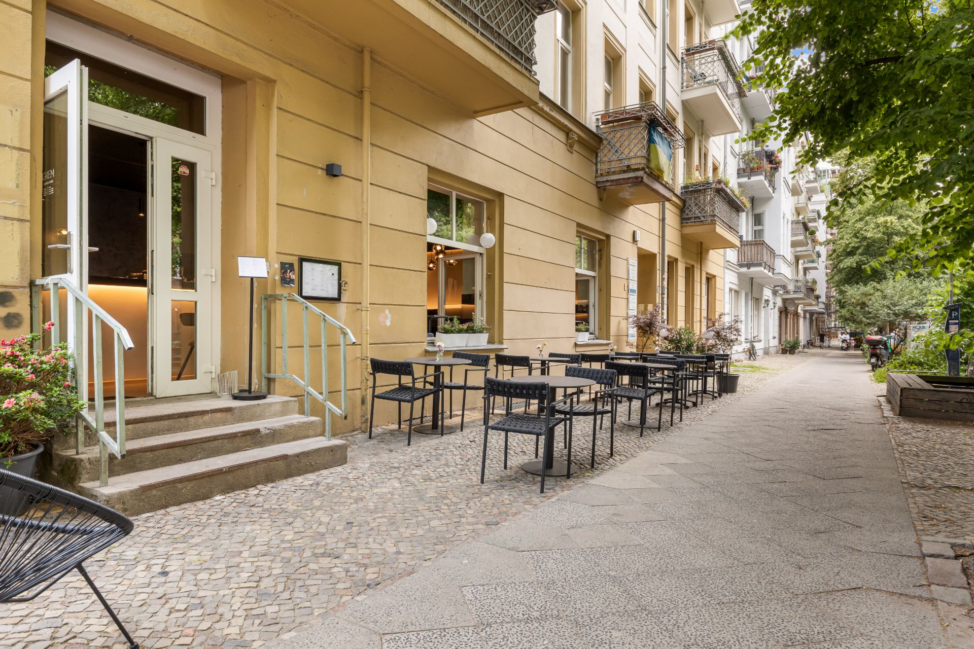 Mehrere Tische und Stühle stehen vor einem Wohnhaus auf einem Gehweg.