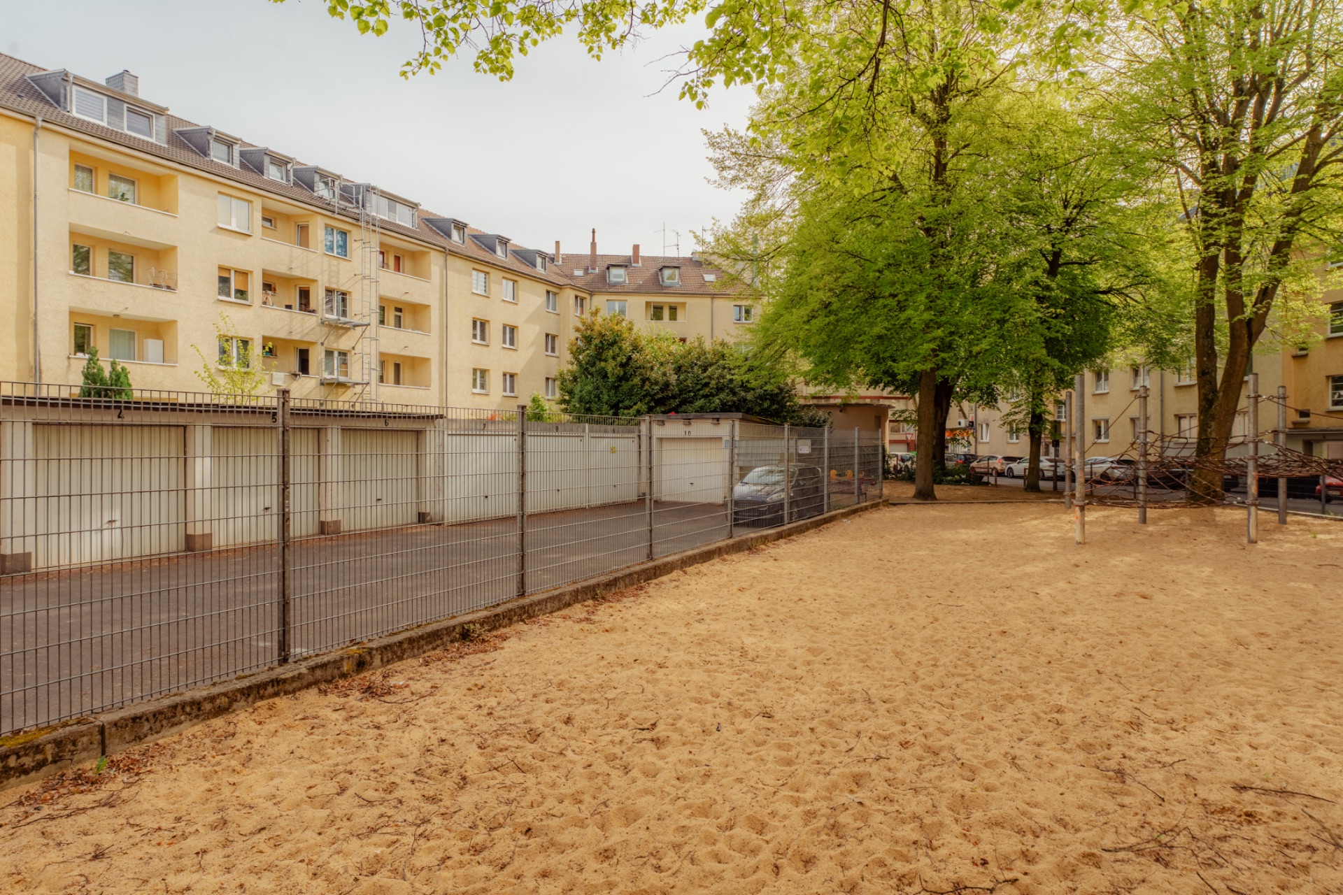 Sandplatz mit Bäumen, Metallzaun und gelben Wohngebäuden im Hintergrund.