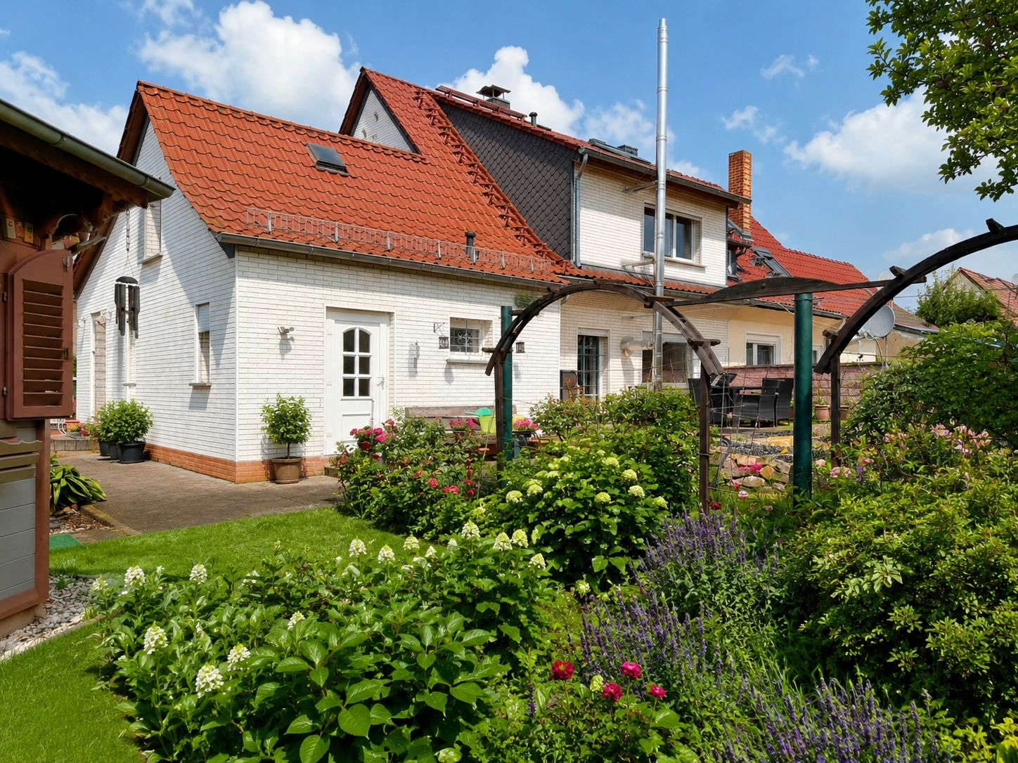 Weißes Wohnhaus mit rotem Dach, umgeben von einem blühenden Garten und Rasen.