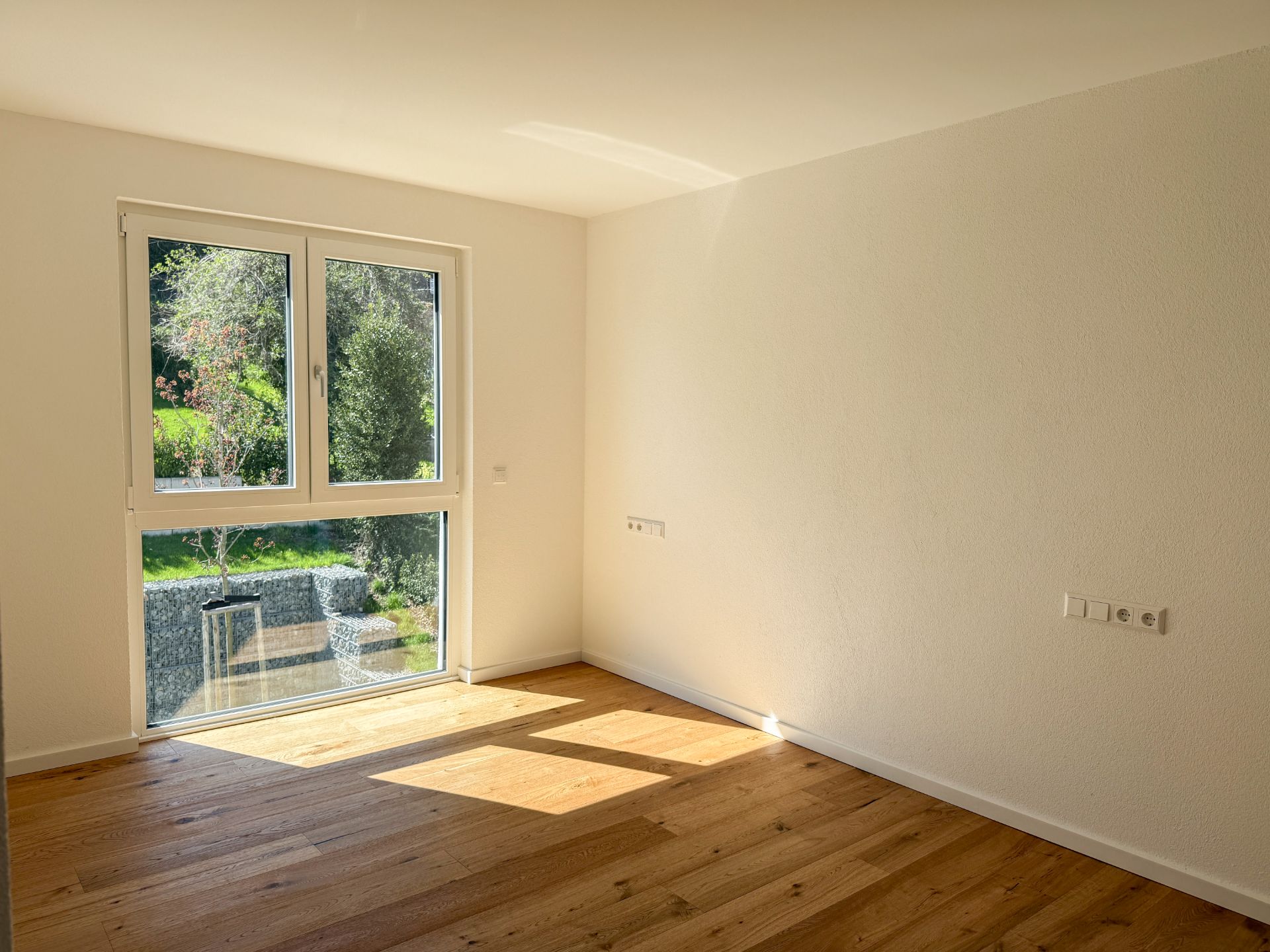 Helles, leeres Zimmer mit großem Fenster und Parkettboden.