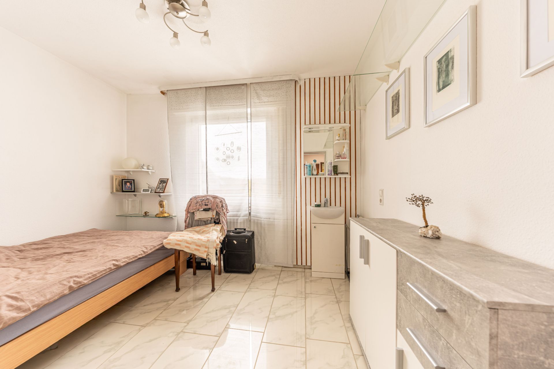 Helles Schlafzimmer mit Bett, Stuhl, Regal, Kommode und Fenster mit Vorhang.