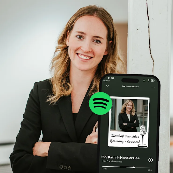 Junge Frau in schwarzem Blazer steht lächelnd an einer weißen Wand, Smartphone mit Spotify-App im Vordergrund.