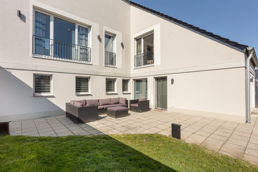 Moderne Terrasse mit Sitzgruppe, Fliesenboden und angrenzendem Rasen vor einem hellen Wohnhaus.