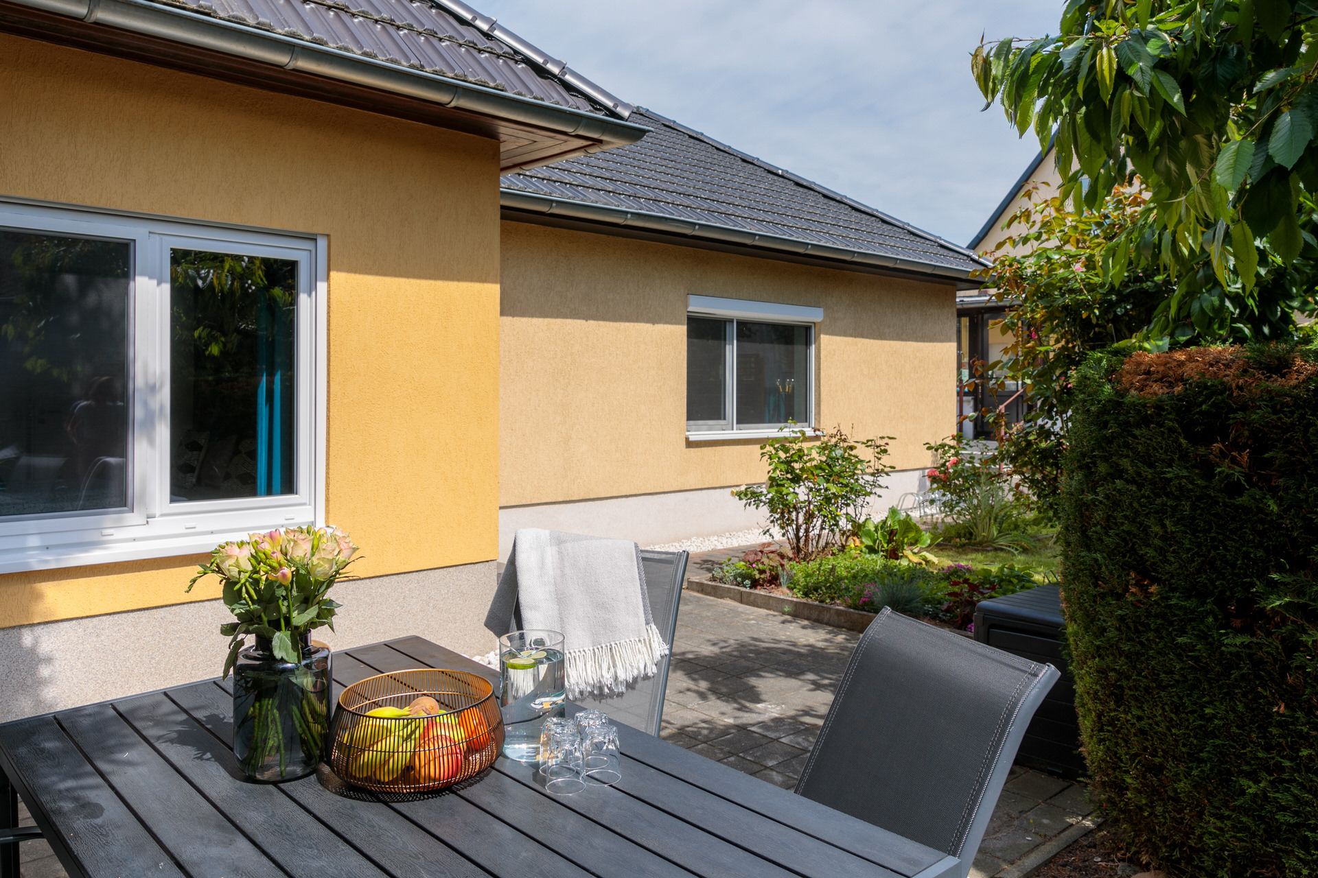 Terrasse mit dunklem Tisch, Stühlen, Obstkorb und Blumen, vor gelbem Haus mit Garten.