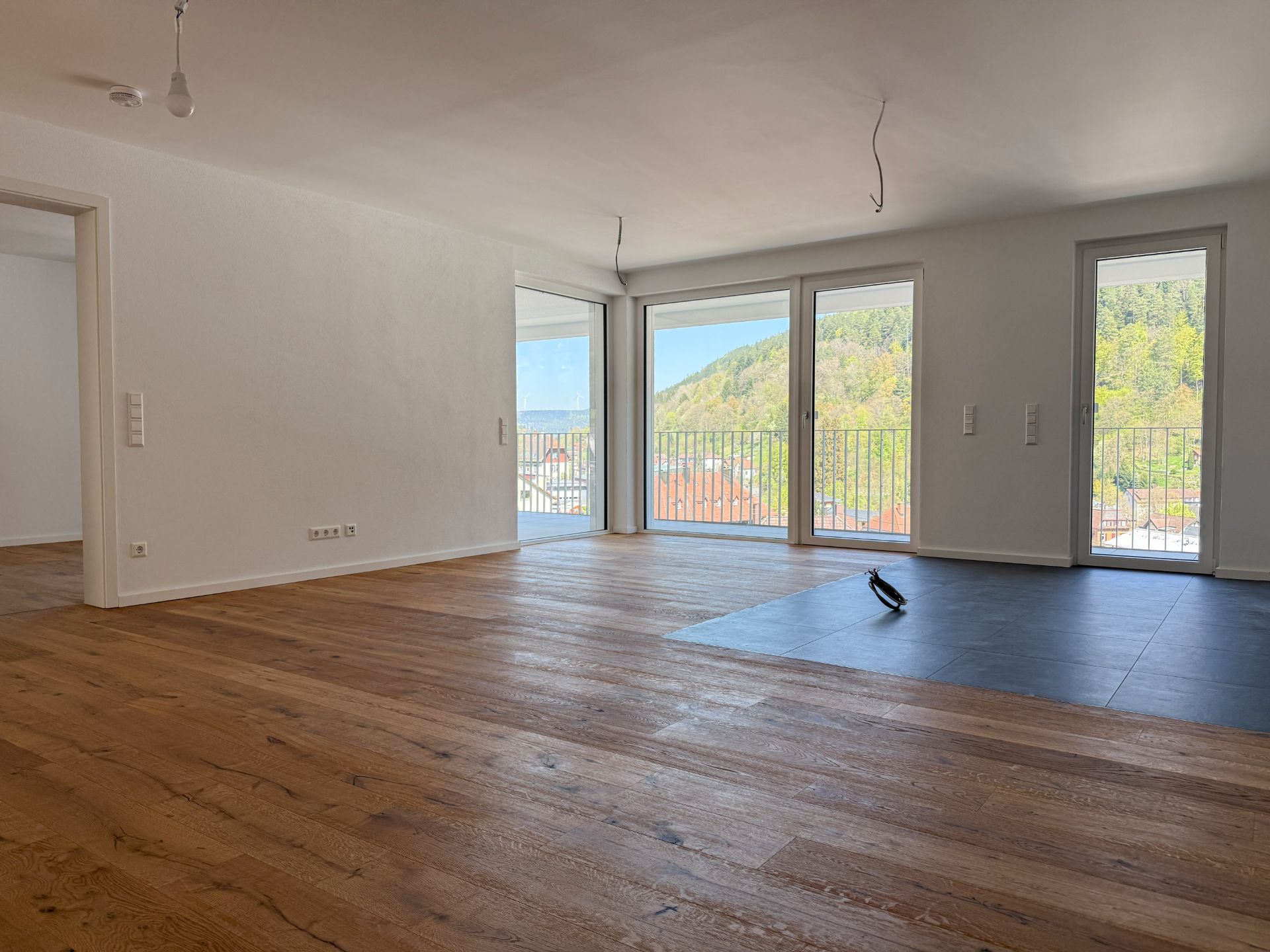 Helles Wohnzimmer mit großen Fenstern, Holzboden und Blick auf einen grünen Hügel.