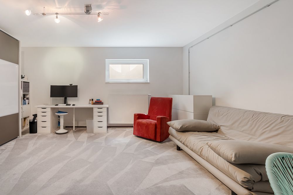 Helles Zimmer mit Schreibtisch, rotem Sessel und grauem Sofa auf hellem Teppich.