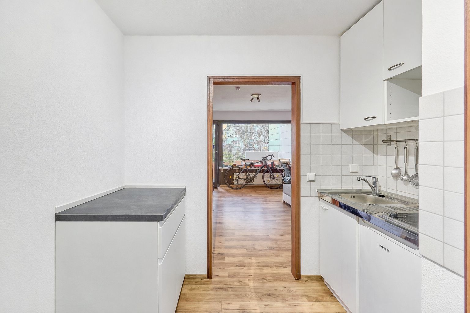 Moderne, kleine Küche mit weißen Schränken und Blick in ein Zimmer mit Fahrrad.