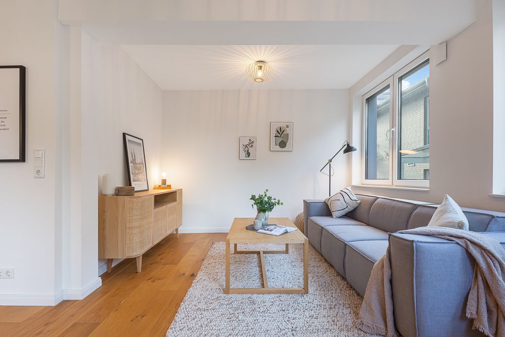 Helles Wohnzimmer mit grauem Sofa, Holztisch, Teppich und großen Fenstern.