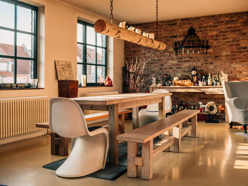 Essbereich mit Holztisch, zwei Bänken, weißen Stühlen und einer freiliegenden Backsteinwand.