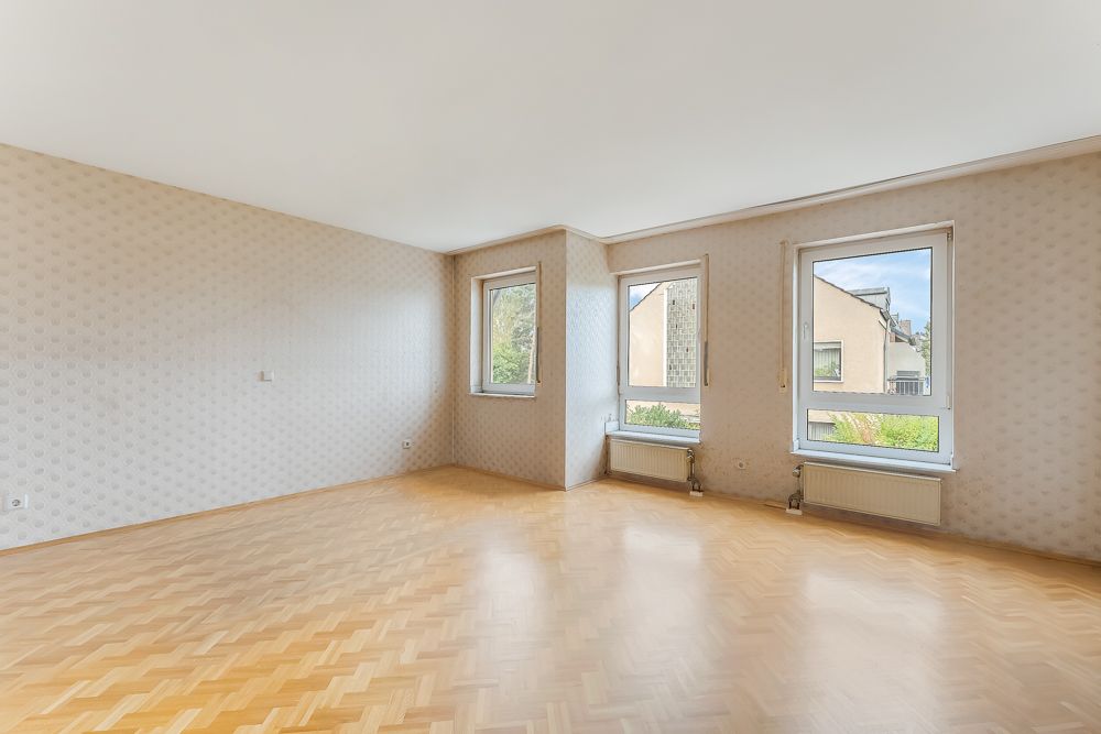 Helles, leeres Zimmer mit Parkettboden, drei Fenstern und gemusterter Tapete.