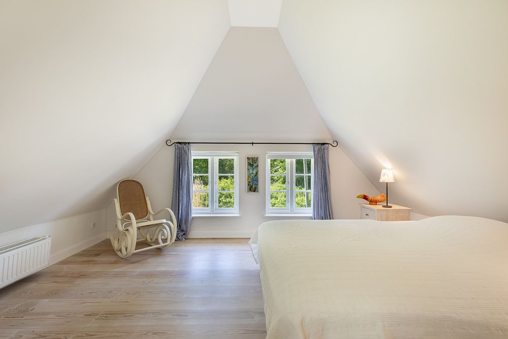 Helles Schlafzimmer mit Dachschräge, Bett, Schaukelstuhl und Fenstern mit Blick ins Grüne.