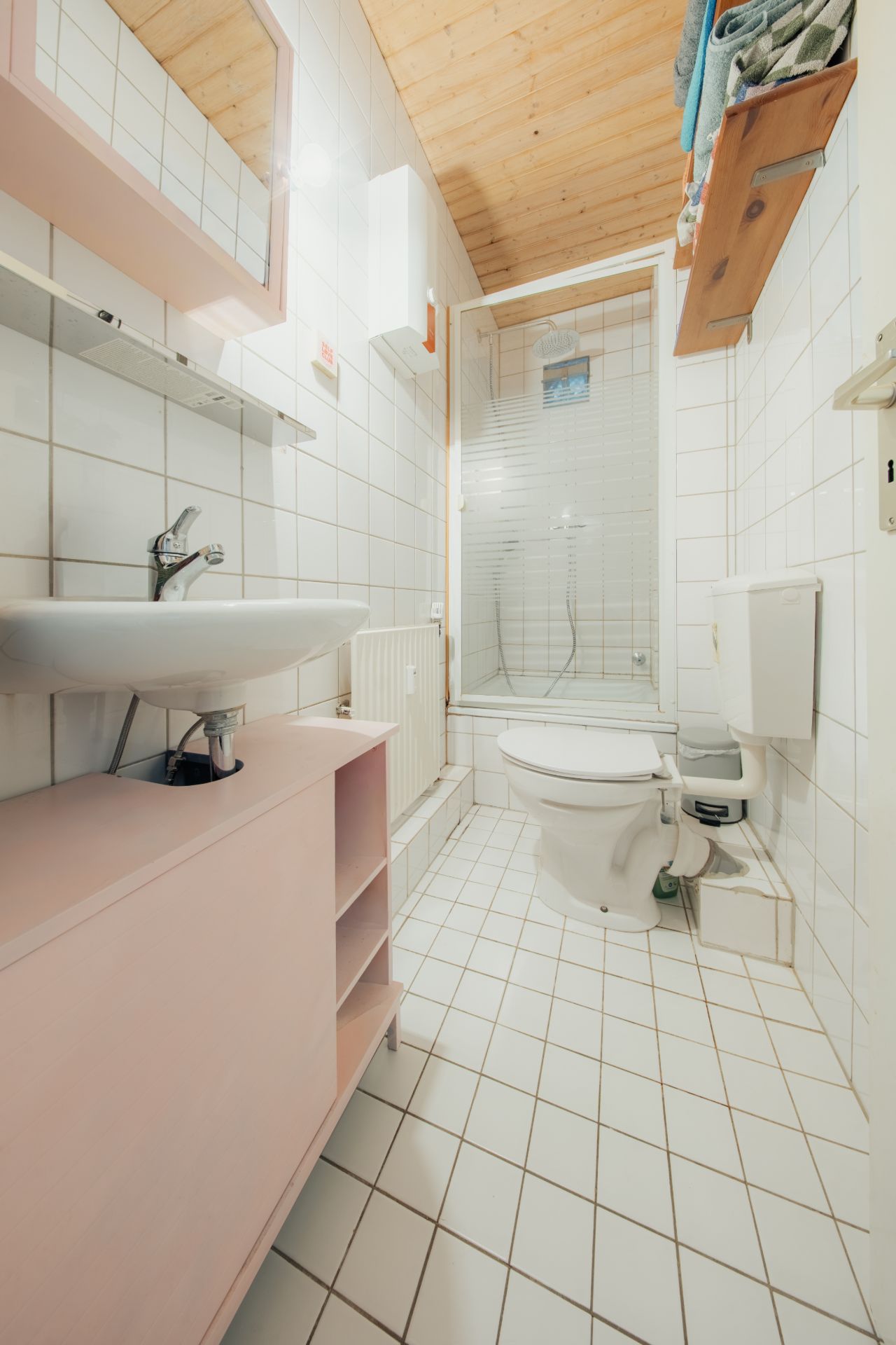 Helles Badezimmer mit Waschbecken, Toilette, Dusche und Regal unter einer Holzdecke.