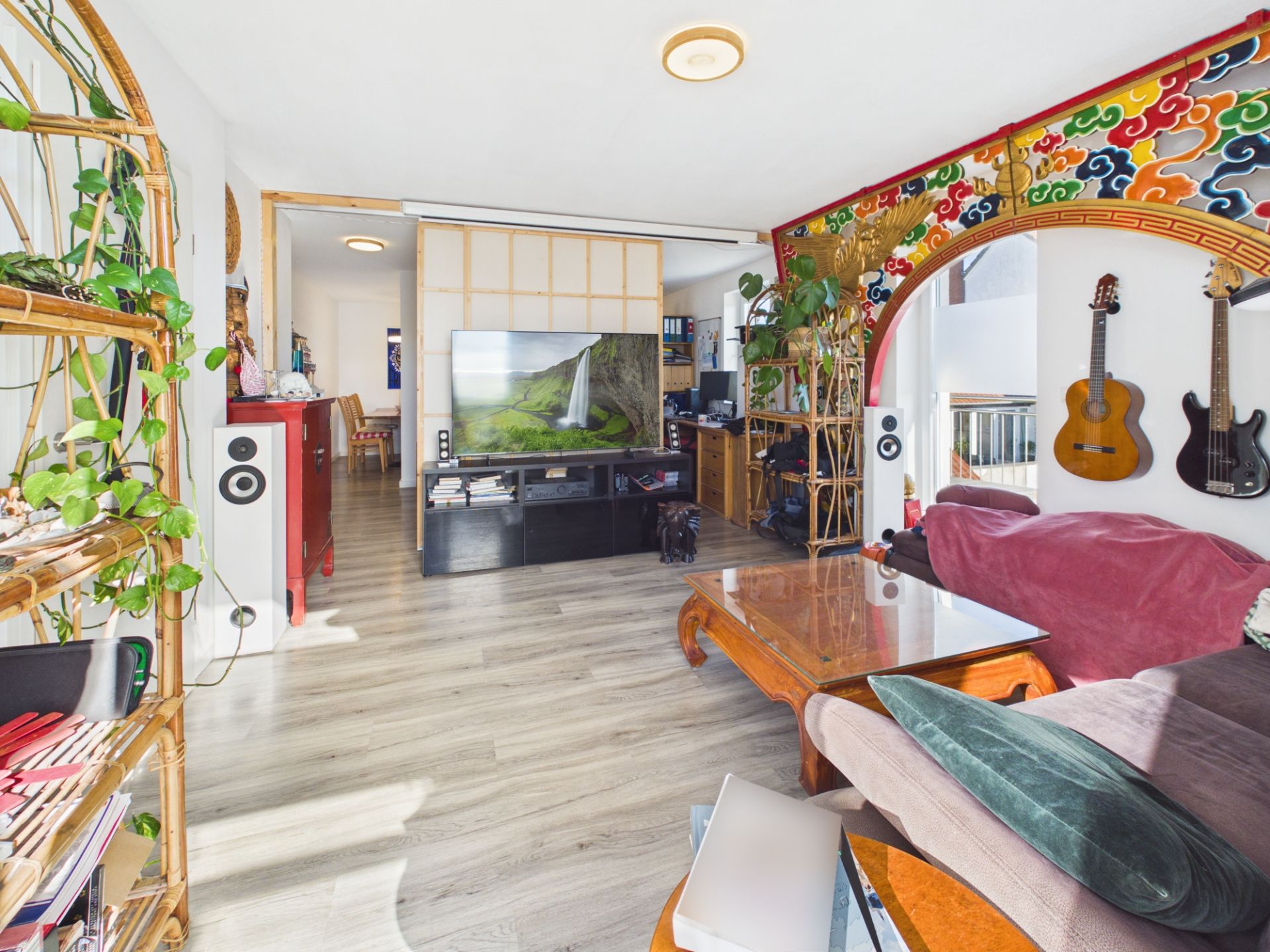 Helles Wohnzimmer mit Couch, Fernseher, Pflanzen, buntem Wandbogen und zwei Gitarren an der Wand.