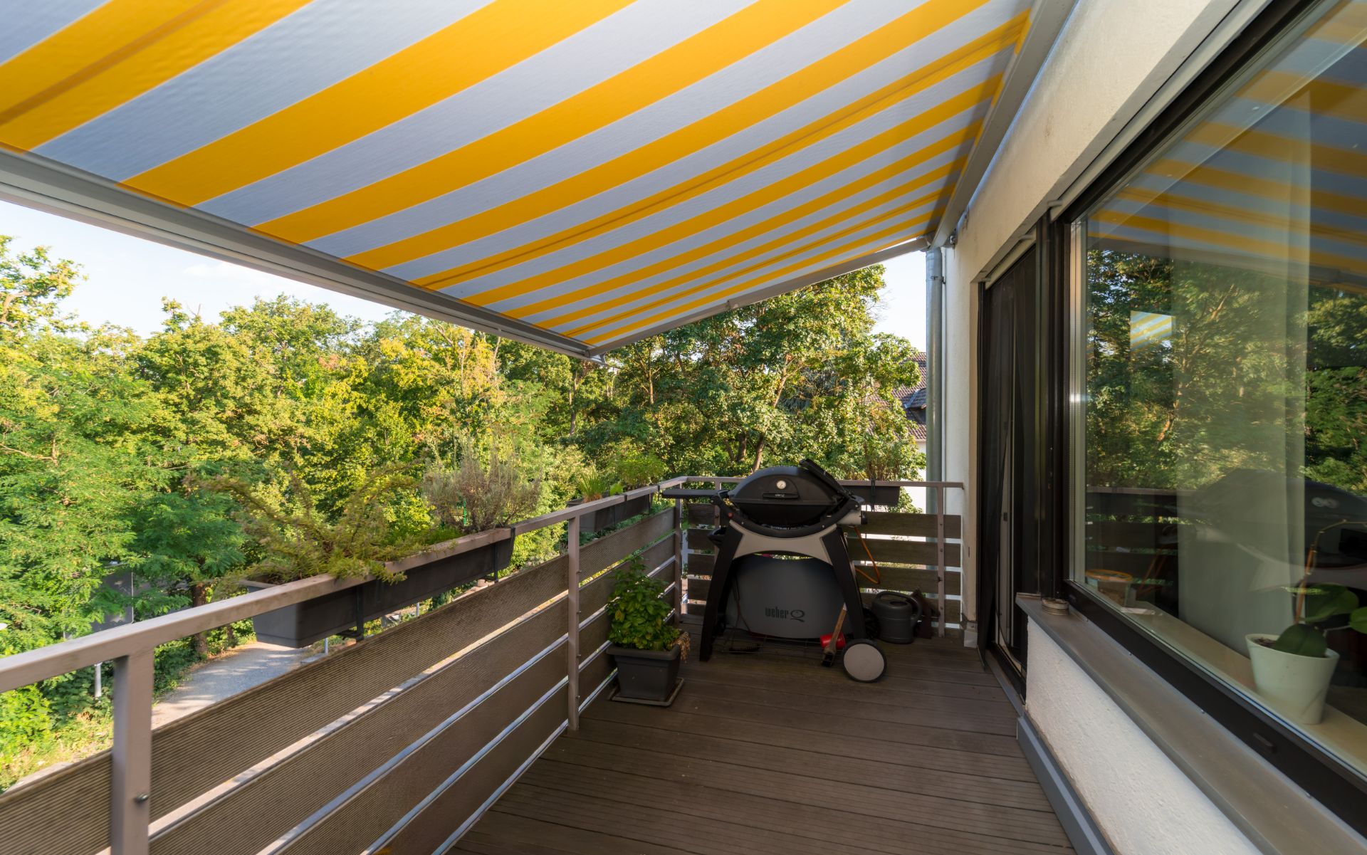 Balkon mit Markise, Grill und Pflanzen, umgeben von Bäumen.