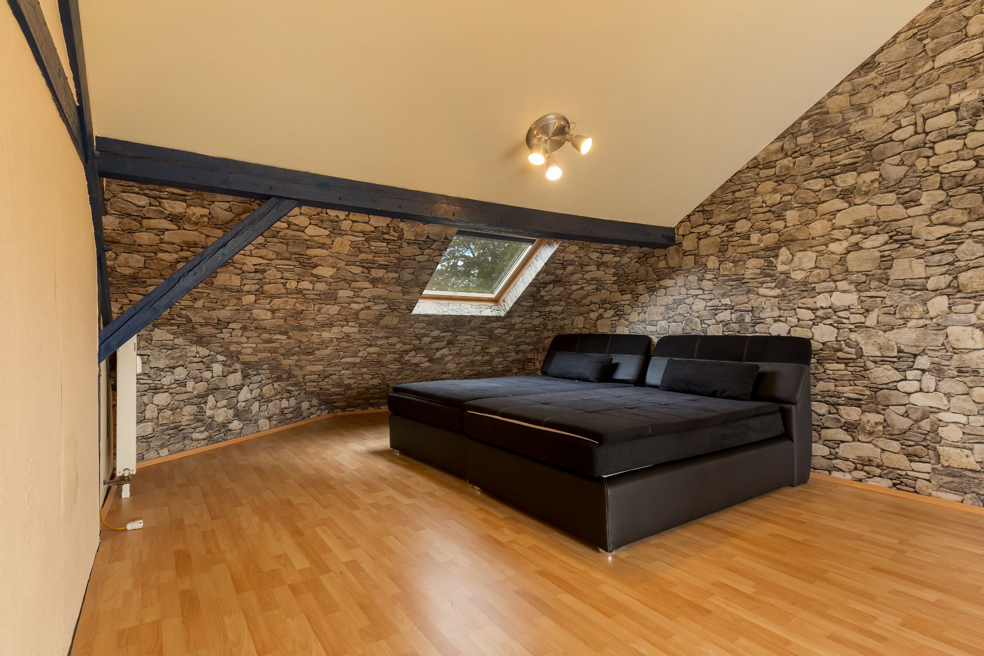 Dachzimmer mit schräger Decke, Steinoptik-Wänden, Dachfenster und schwarzem Sofa auf Laminatboden.