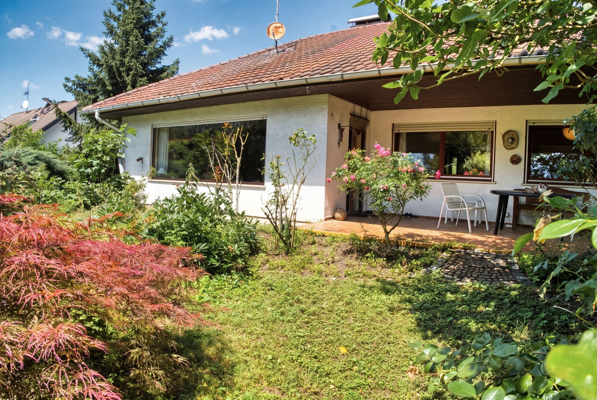 Einfamilienhaus mit Terrasse und Garten, umgeben von grünen Pflanzen und Sträuchern.