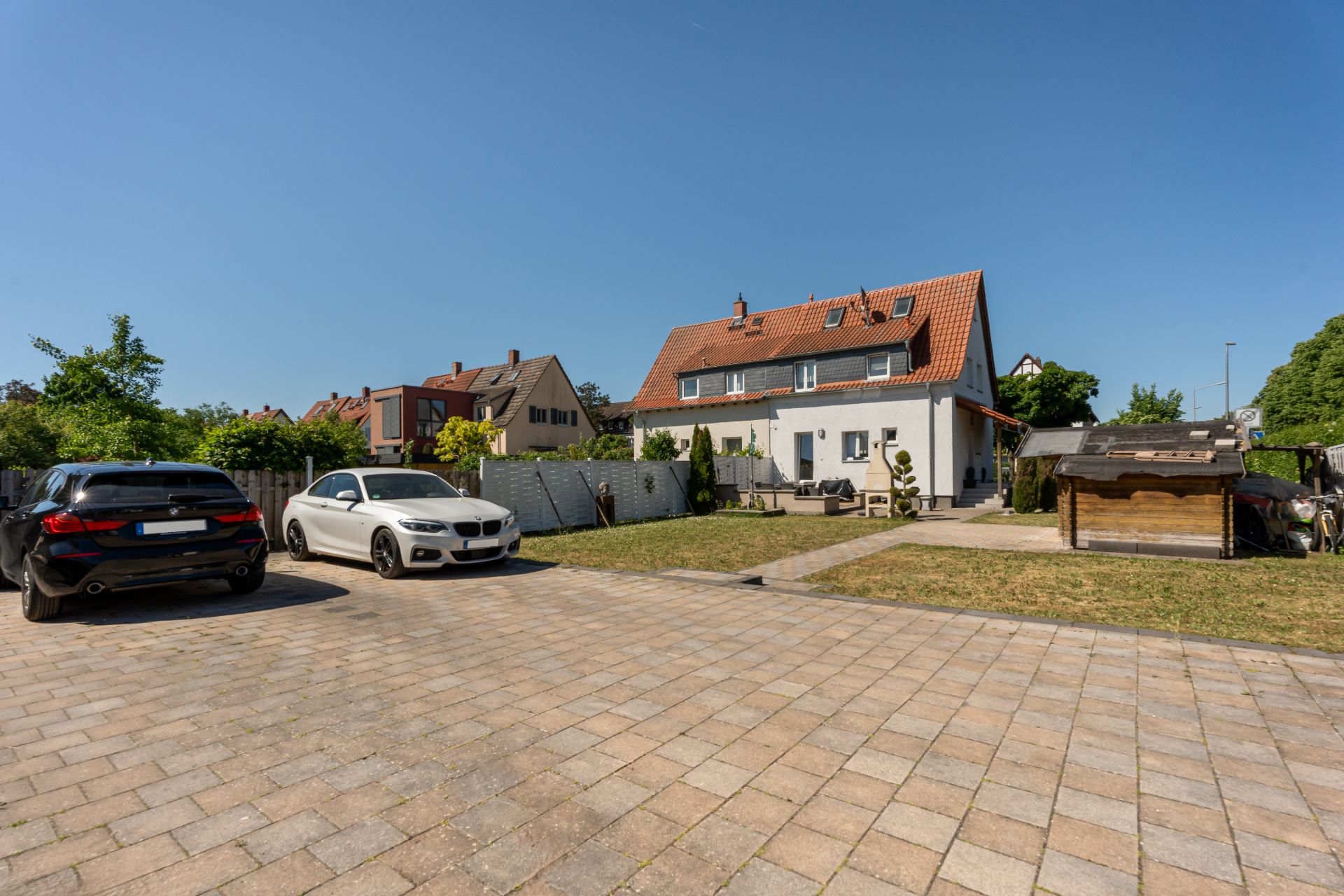 Doppelhaushälfte mit Garten, gepflastertem Parkplatz und zwei Autos im Vordergrund.