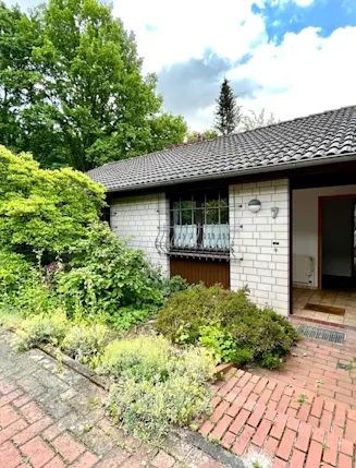 Eingang eines Hauses mit hellem Mauerwerk, roten Ziegelsteinen und umgeben von Sträuchern.