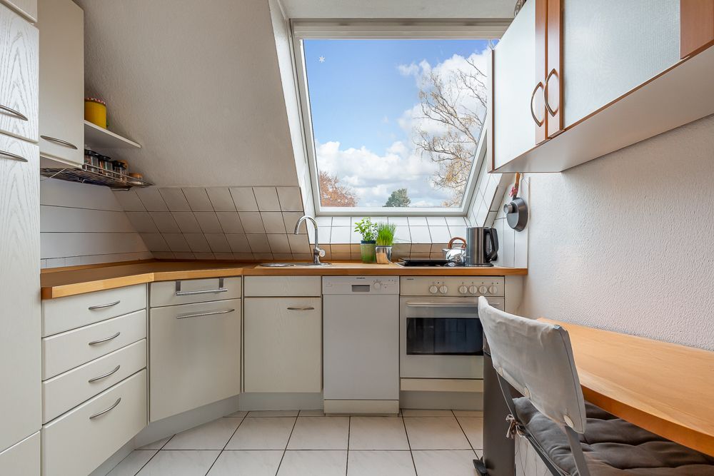 Helle Einbauküche mit großem Fenster, Herd, Spüle und weißen Schränken unter Dachschräge.